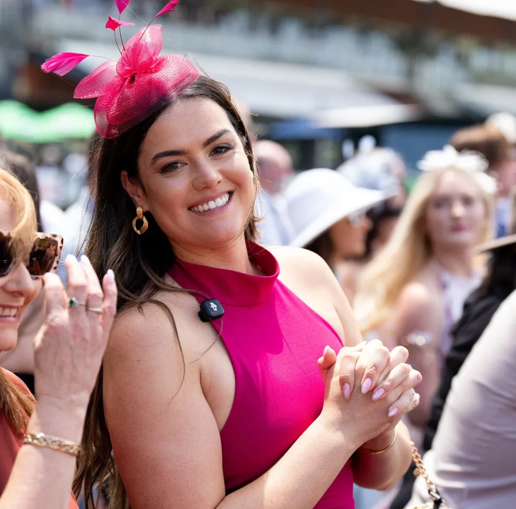 A woman in a pink dress and fascinator smiles in the crowd at the races.