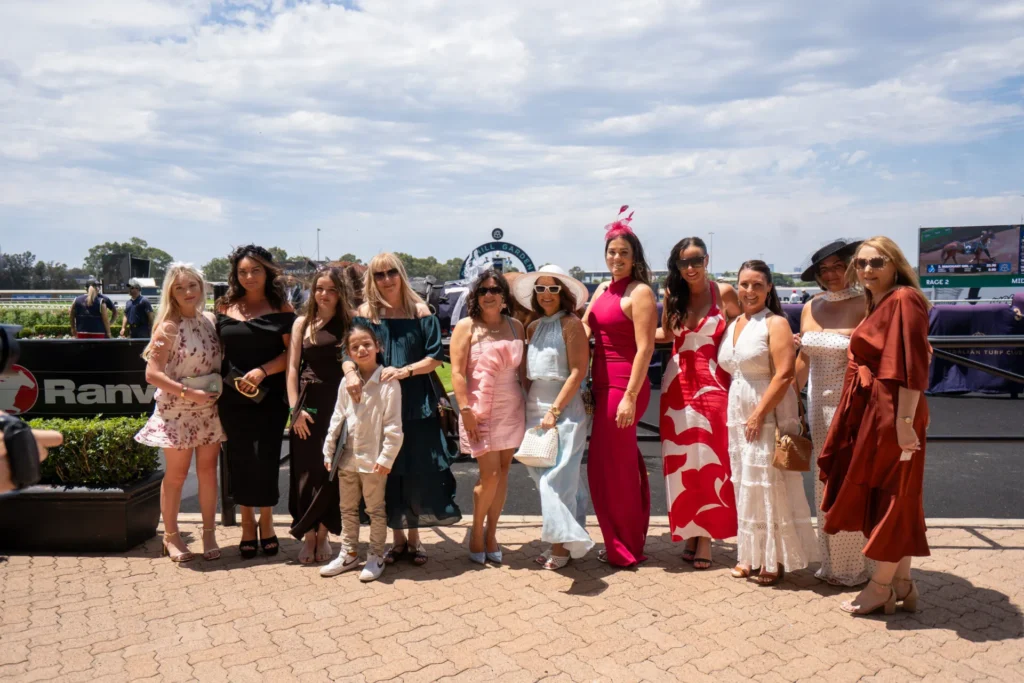 A group of women in race-day dresses stand together beside the track.